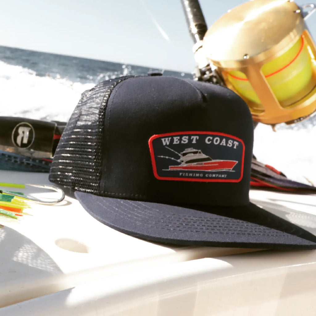 A black trucker-style cap with a yellow and white 'West Coast Fishing Company' patch on the front, photographed on a boat with fishing gear and ocean water in the background.